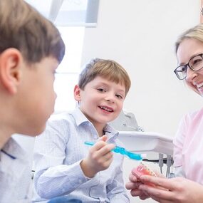 Zahnärztin zeigt zwei Jungen spielerisch das Zähneputzen mit einem Gebissmodell.